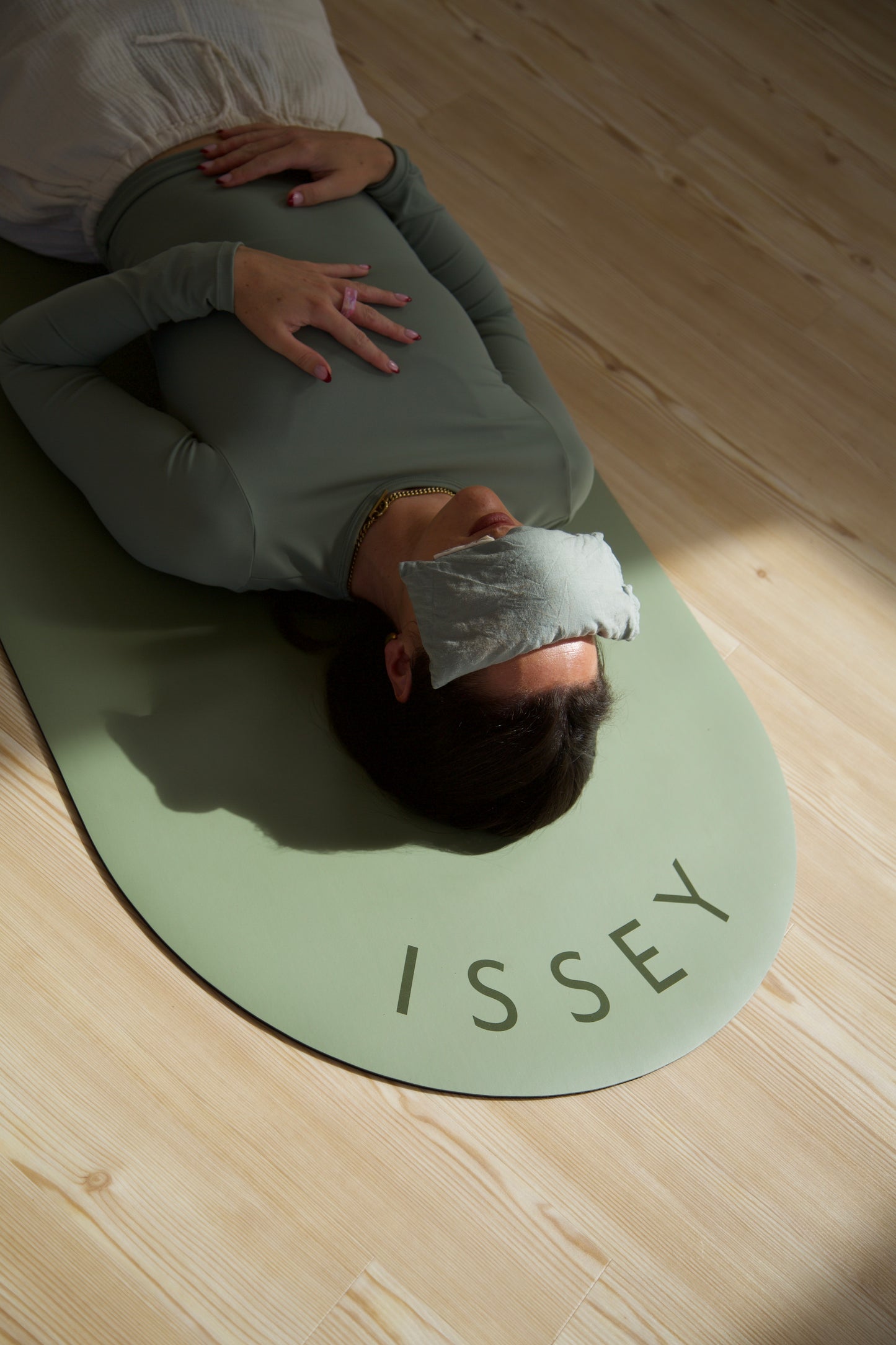 A person laying on a green eco yoga mat with the brand name 'ISSEY' printed on it, resting with an eyepillow.