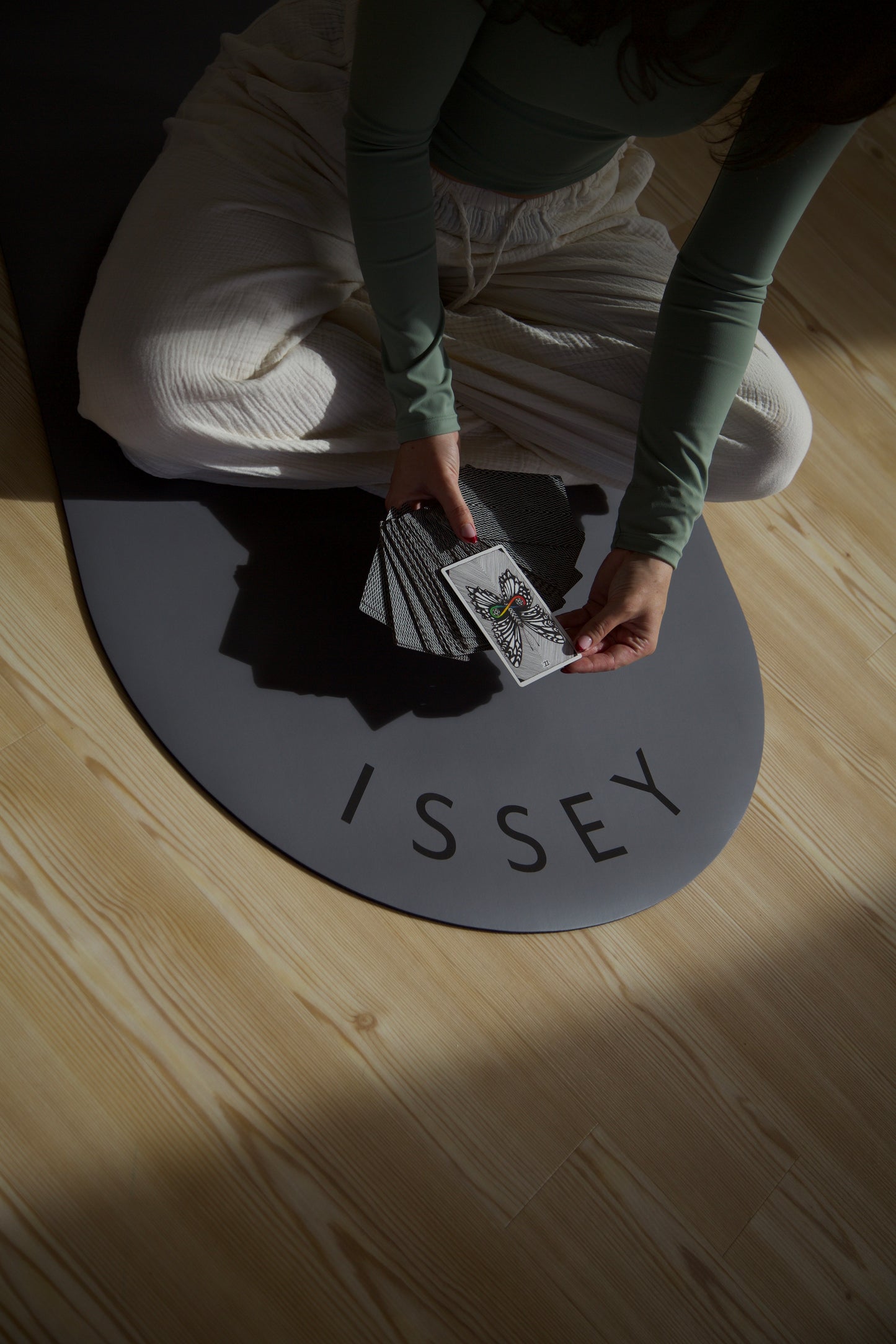 A person sitting on a dark gray eco yoga mat with the brand name 'ISSEY' printed on it, holding a tarot card.